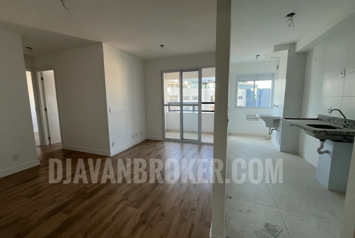 Foto do interior de um apartamento residencial vazio e recém-entregue, mostrando a sala de estar e a cozinha integrada, conforme o arquivo Sala e Cozinha. À esquerda, a sala possui piso laminado imitando madeira em tom médio, paredes brancas e uma porta de correr de vidro de três folhas que dá acesso à varanda. À direita, a cozinha e a área de serviço possuem piso frio em cerâmica clara, paredes revestidas com azulejos brancos, uma bancada de granito escuro com pia e um tanque de lavar roupas ao fundo, próximo a uma janela de alumínio branco. O teto de gesso branco exibe fiações expostas prontas para a instalação de luminárias. A marca d'água "DJAVANBROKER.COM" está centralizada na imagem em letras semitransparentes.