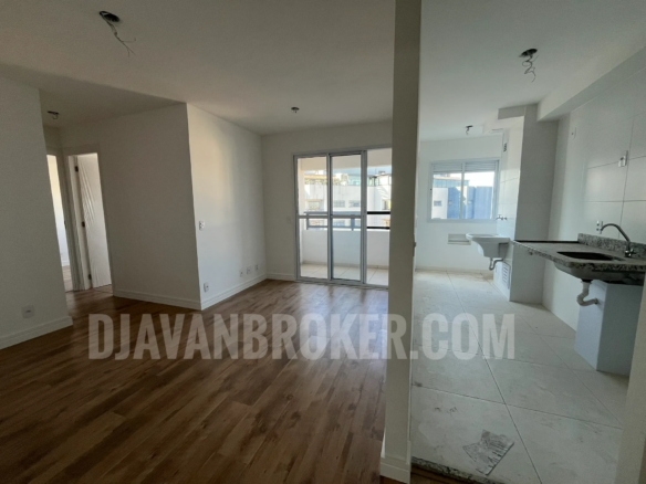 Foto do interior de um apartamento residencial vazio e recém-entregue, mostrando a sala de estar e a cozinha integrada, conforme o arquivo Sala e Cozinha. À esquerda, a sala possui piso laminado imitando madeira em tom médio, paredes brancas e uma porta de correr de vidro de três folhas que dá acesso à varanda. À direita, a cozinha e a área de serviço possuem piso frio em cerâmica clara, paredes revestidas com azulejos brancos, uma bancada de granito escuro com pia e um tanque de lavar roupas ao fundo, próximo a uma janela de alumínio branco. O teto de gesso branco exibe fiações expostas prontas para a instalação de luminárias. A marca d'água "DJAVANBROKER.COM" está centralizada na imagem em letras semitransparentes.