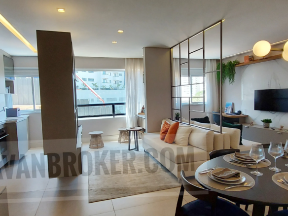 Uma vista panorâmica interna de conceito aberto de um apartamento moderno na Vila Rosália, Guarulhos, conforme mostrado no arquivo Salas_cozinha e varanda_Vila_Rosalia. No primeiro plano à direita, uma mesa de jantar redonda e escura está elegantemente posta para seis pessoas com copos de cristal. Na frente dela, uma sala de estar sofisticada com um sofá creme e uma divisória de metal decorativa com plantas penduradas. Uma TV de tela plana está montada em um painel de parede de mármore branco com iluminação de fundo. À esquerda, uma cozinha moderna com armários em tons de cinza, geladeira, cooktop... Ao fundo, uma varanda envidraçada com tamboretes de design e vista urbana. Uma marca d'água 'DJAVANBROKER.COM' está visível no centro da imagem.