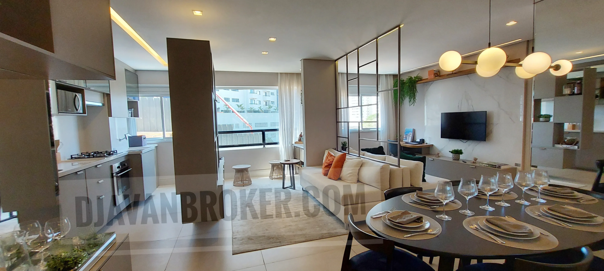 Uma vista panorâmica interna de conceito aberto de um apartamento moderno na Vila Rosália, Guarulhos, conforme mostrado no arquivo Salas_cozinha e varanda_Vila_Rosalia. No primeiro plano à direita, uma mesa de jantar redonda e escura está elegantemente posta para seis pessoas com copos de cristal. Na frente dela, uma sala de estar sofisticada com um sofá creme e uma divisória de metal decorativa com plantas penduradas. Uma TV de tela plana está montada em um painel de parede de mármore branco com iluminação de fundo. À esquerda, uma cozinha moderna com armários em tons de cinza, geladeira, cooktop... Ao fundo, uma varanda envidraçada com tamboretes de design e vista urbana. Uma marca d'água 'DJAVANBROKER.COM' está visível no centro da imagem.