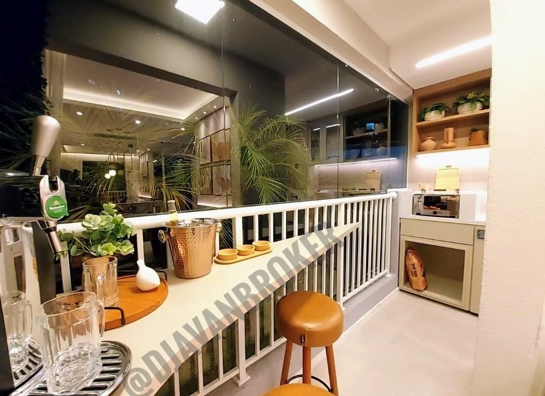 Varando do apartamento de 43m². A varanda está fechada por vidro e possui uma churrasqueira a carvão ecológica, guarda corpo de aço, no lado esquerdo tem dois bancos e sobre estes bancos consta uma mesa com vasos e uma chopeira Heineken.